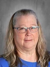 Woman with glasses and long hair, wearing a blue shirt, against a gray background.