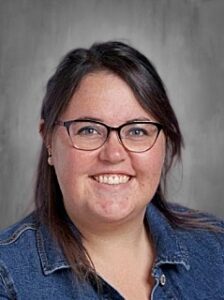 Smiling woman with glasses and dark hair, wearing a denim jacket, against a gray background.