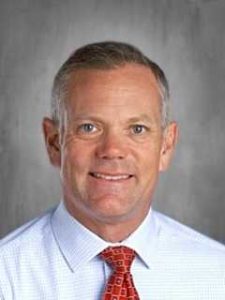 Man in a light blue shirt and red tie, smiling, with short hair, against a gray background.