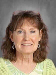 Smiling woman with curly brown hair, wearing green attire and earrings, against a gray background.