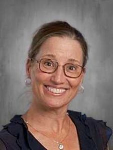 Smiling woman with glasses and a dark top, set against a gray background.