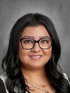 A person with long dark hair and glasses smiles at the camera against a gray background.