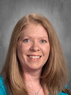 Smiling woman with long, straight brown hair wearing a blue top, set against a neutral background.