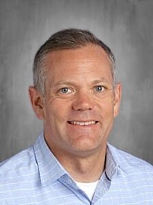 Headshot of a smiling man with short gray hair, wearing a light blue checkered shirt against a neutral background.