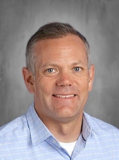 Headshot of a smiling man with short gray hair, wearing a light blue checkered shirt against a neutral background.
