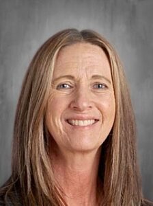 Portrait of a woman with long brown hair and a warm smile, set against a gray background.