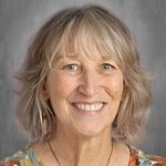 A smiling woman with medium-length wavy hair, wearing a colorful floral shirt, against a gray background.