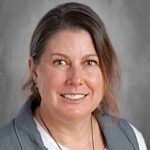 Smiling woman with shoulder-length brown hair, wearing a gray vest over a white shirt, against a neutral background.