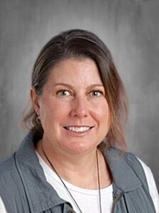 Smiling woman with shoulder-length brown hair, wearing a gray vest over a white shirt, against a neutral background.