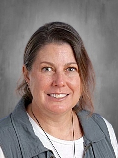 Smiling woman with shoulder-length brown hair, wearing a gray vest over a white shirt, against a neutral background.