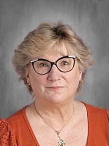 A woman with short, wavy blonde hair and glasses, wearing an orange blouse, poses for a professional portrait.