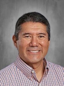 A smiling man with short dark hair, wearing a checkered shirt, poses against a gray background.