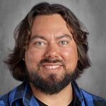 A smiling man with long, dark hair and a beard, wearing a blue plaid shirt against a gray background.