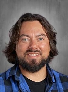 A smiling man with long, dark hair and a beard, wearing a blue plaid shirt against a gray background.