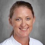 Headshot of a woman with light brown hair, smiling, wearing a white shirt, with a gray background.