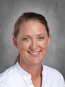 Headshot of a woman with light brown hair, smiling, wearing a white shirt, with a gray background.