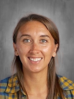 Smiling woman with long brown hair, wearing a plaid shirt, against a gray background.