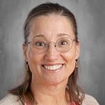 Smiling woman with glasses and long hair, wearing a pink shirt and beige cardigan, set against a gray background.