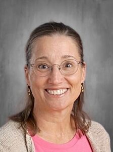 Smiling woman with glasses and long hair, wearing a pink shirt and beige cardigan, set against a gray background.