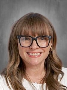 A smiling woman with long, wavy hair and glasses poses against a gray background.