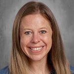 A woman with long, light brown hair smiles at the camera, wearing a striped shirt and denim jacket.