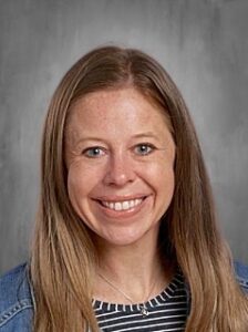 A woman with long, light brown hair smiles at the camera, wearing a striped shirt and denim jacket.