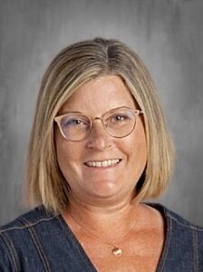 A woman with shoulder-length blonde hair and glasses smiles warmly, wearing a denim top against a gray background.