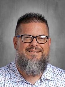 A smiling man with glasses and a beard, wearing a patterned shirt, against a gray background.