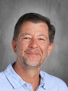 A smiling middle-aged man with short hair and a beard, wearing a light blue collared shirt.
