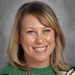 Smiling woman with shoulder-length blonde hair wearing a green sweater with a white pattern against a gray background.