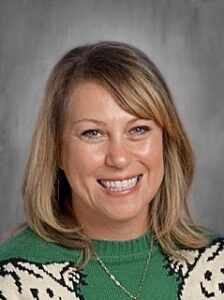 Smiling woman with shoulder-length blonde hair wearing a green sweater with a white pattern against a gray background.