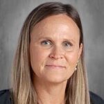 Portrait of a woman with long brown hair, wearing a black shirt and pearl necklace, against a gray background.