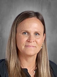 Portrait of a woman with long brown hair, wearing a black shirt and pearl necklace, against a gray background.