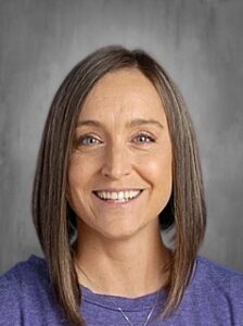 Smiling woman with shoulder-length brown hair, wearing a purple shirt, against a gray background.