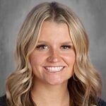 Smiling woman with long, wavy blonde hair, wearing a black top, against a gray background.