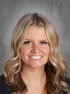 Smiling woman with long, wavy blonde hair, wearing a black top, against a gray background.
