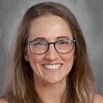 Smiling woman with long brown hair and glasses, wearing a black top, against a gray background.