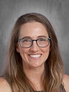 Smiling woman with long brown hair and glasses, wearing a black top, against a gray background.