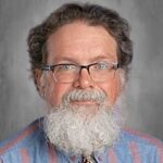 A middle-aged man with glasses and a full beard, wearing a blue shirt with stripes and a patterned tie.