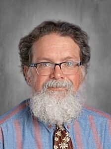 A middle-aged man with glasses and a full beard, wearing a blue shirt with stripes and a patterned tie.