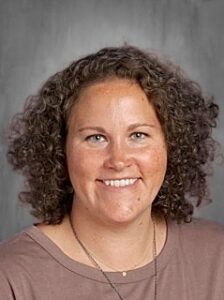 Smiling woman with curly hair, wearing a brown top, against a neutral gray background.