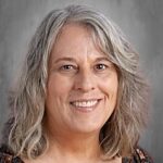 A smiling woman with wavy gray hair wearing a patterned top, against a neutral background.