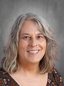 A smiling woman with wavy gray hair wearing a patterned top, against a neutral background.