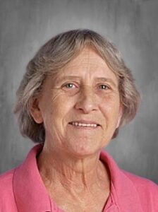 Older woman with short gray hair, smiling, wearing a pink polo shirt, against a gray background.