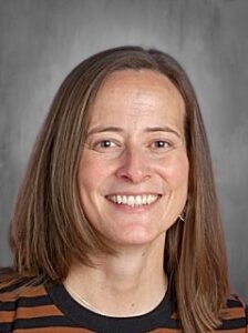 Smiling woman with medium-length brown hair, wearing a striped black and orange shirt, against a gray background.