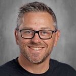 Smiling man with short, gray hair and glasses, wearing a black shirt, against a gray background.