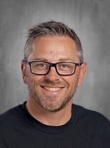 Smiling man with short, gray hair and glasses, wearing a black shirt, against a gray background.