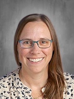 Smiling woman with long brown hair and glasses, wearing a patterned blouse, against a gray background.