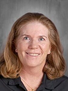 Smiling woman with shoulder-length, wavy hair, wearing a black shirt, against a gray background.