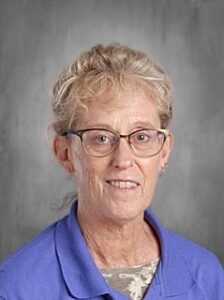 A woman with curly blonde hair and glasses smiles, wearing a blue shirt against a gray background.